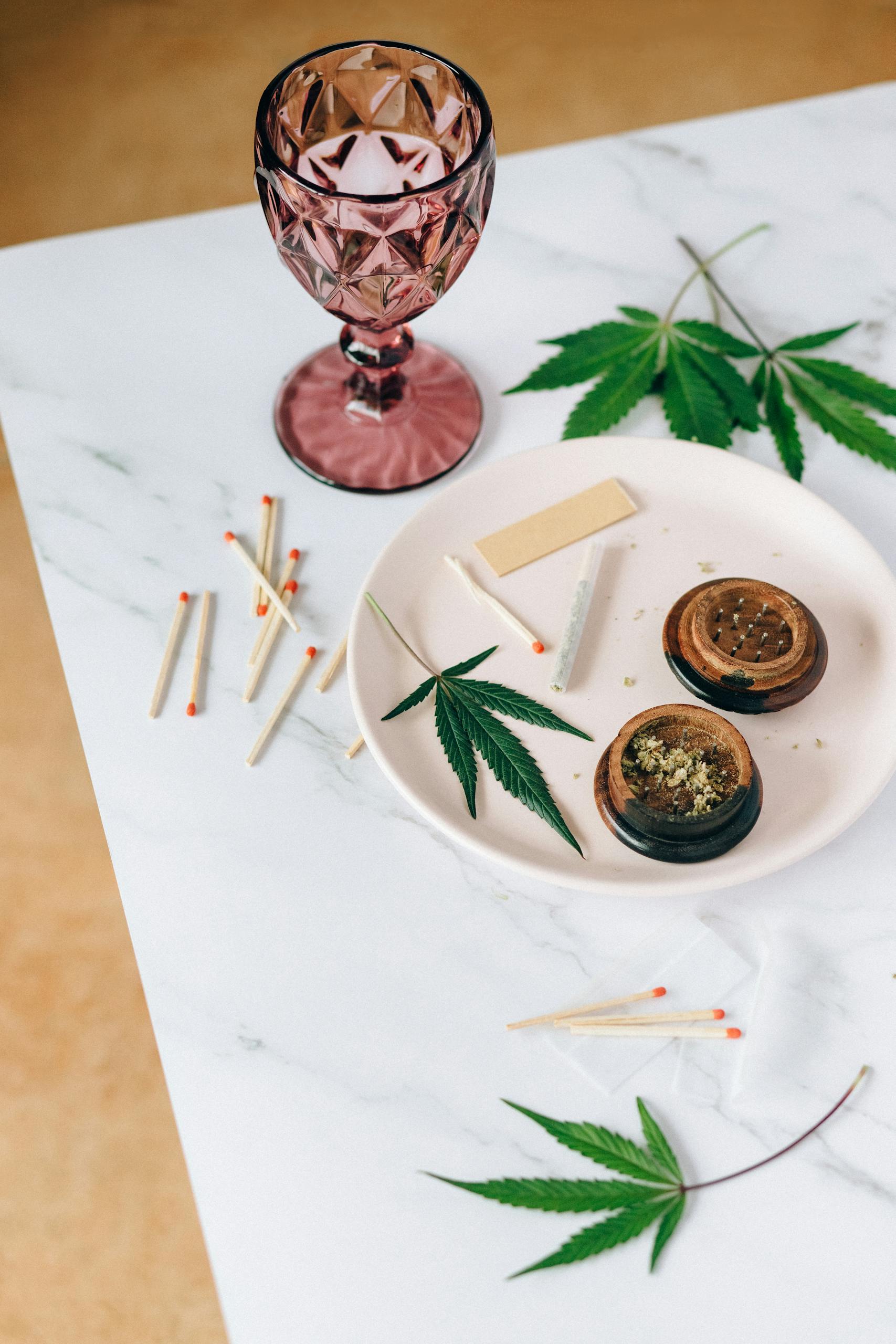 Aesthetic arrangement of cannabis leaves, grinder, and glass of wine on marble surface.